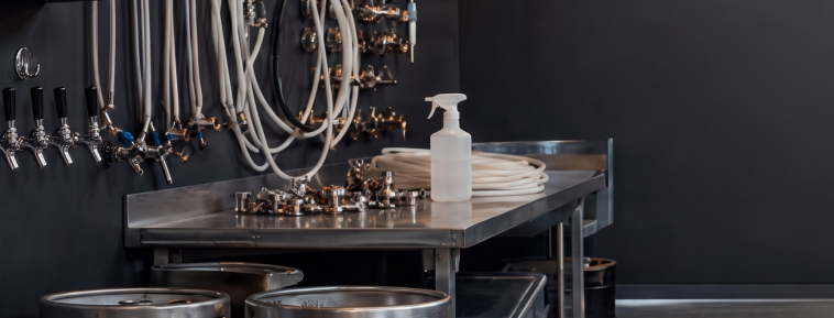 Sanitizing spray bottle staged on a stainless steel worktable in a brewery keg room with beer lines and taps